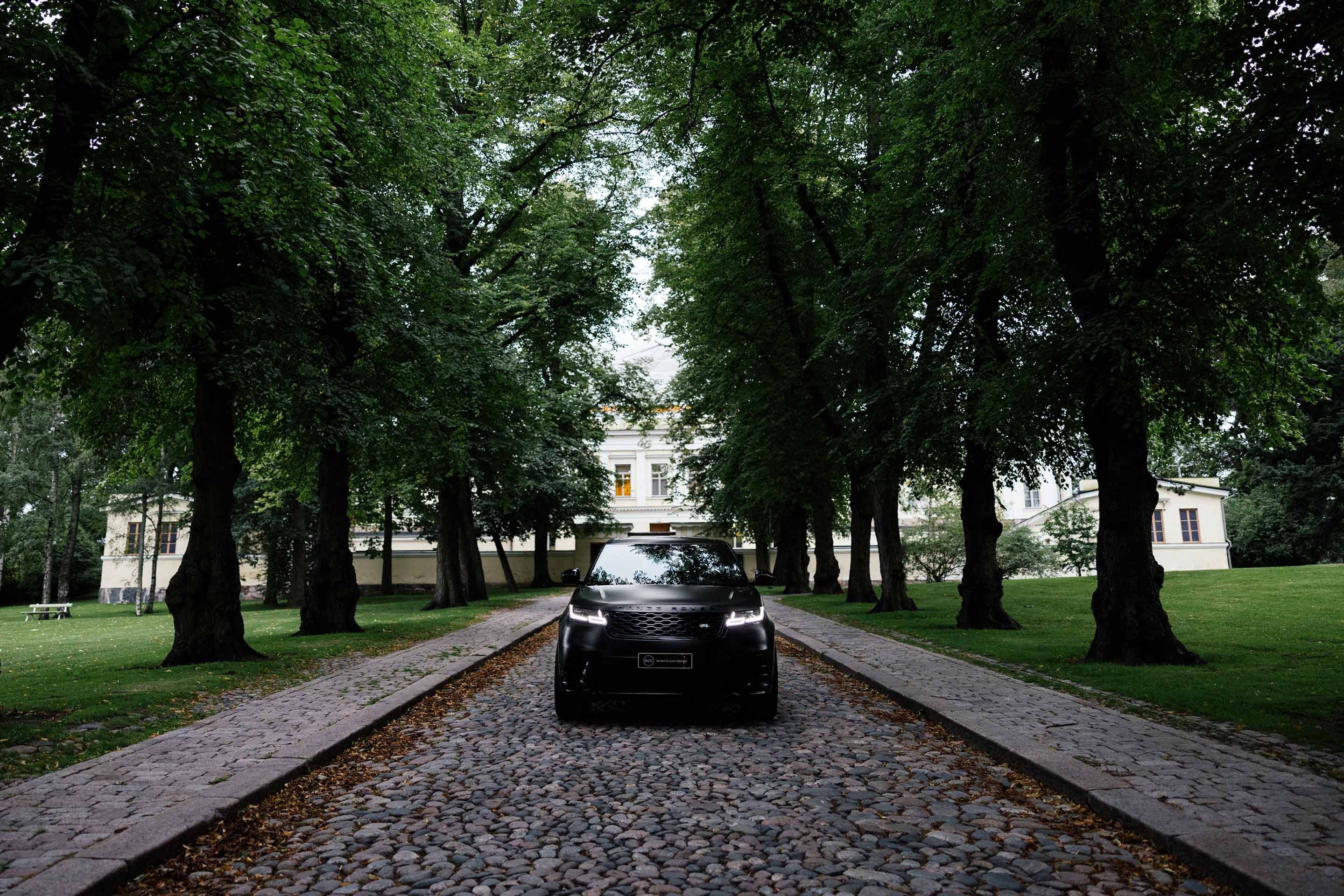 a black range rover velar is parked on a cobblestone driveway surrounded by trees .