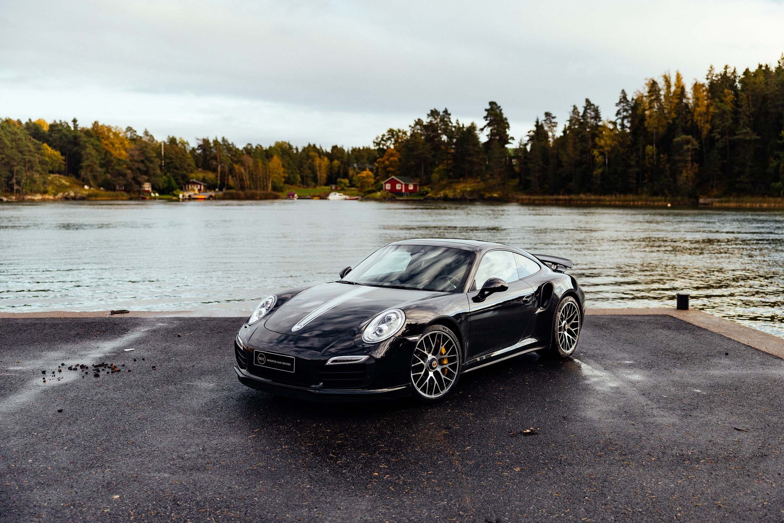 a black porsche 911 turbo s is parked on a dock next to a body of water .